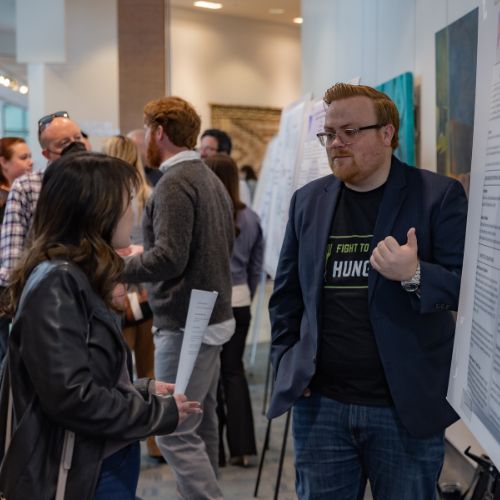 Male student in front of poster speaking to a female attendee.